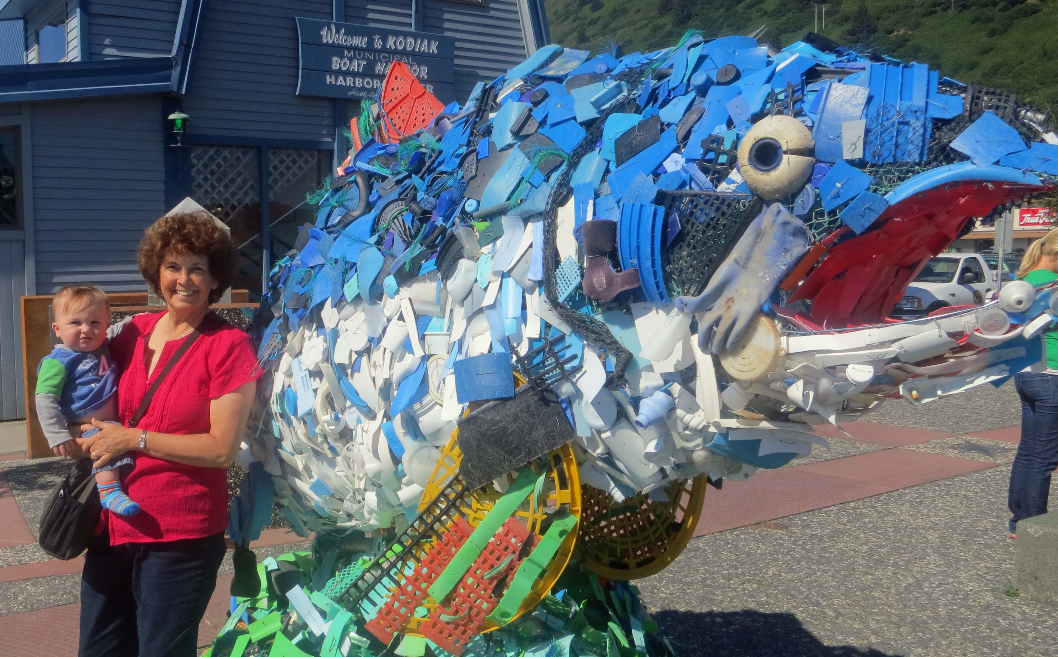 Peggy poses with out youngest grandson, Cooper in front of the Harbor Masters office in Kodiak. The large fish is a sculpture made from trash collected from the ocean. Hopefully Cooper will grow up in a world with less trash.